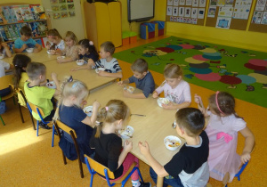 Grupa dzieci siedzi przy stole i je przygotowany deser.